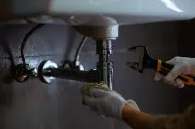 A person in white gloves uses a wrench to work on pipes under a sink
